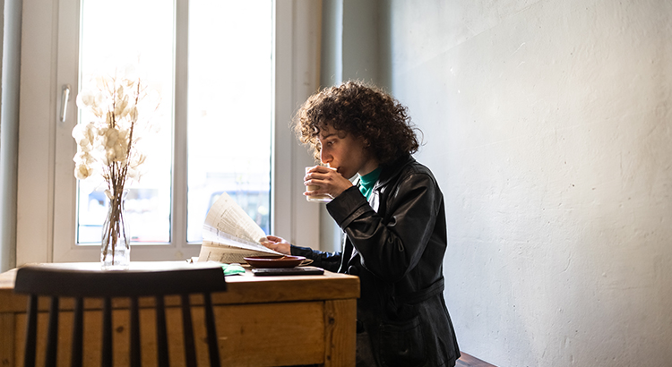 Sipping at a tasty warm coffee, a non-binary young adult sits while reading the newspaper